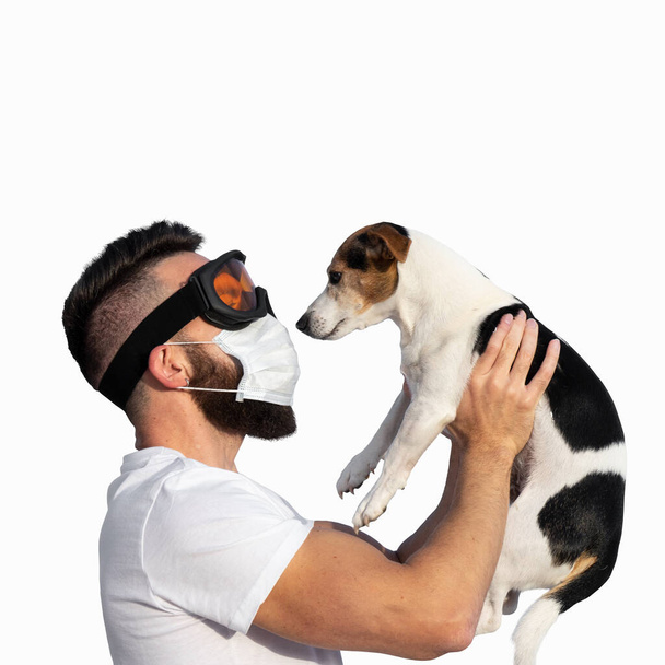 Καυκάσιος γενειοφόρος άνδρας με μάσκα προσώπου και προστατευτικά γυαλιά που κρατούν ψηλά τον χαριτωμένο σκύλο Τζακ Ράσελ κοντά στο πρόσωπο. Έννοια της αλλεργίας κατοικίδιων ζώων ή μυρωδιά σκύλου στο σπίτι, απομονώνονται σε λευκό - Φωτογραφία, εικόνα