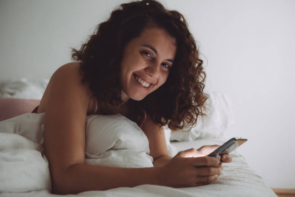 jeune jolie femme avec les cheveux bouclés au lit avec téléphone style de vie domestique
 - Photo, image