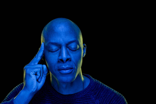 Studio foto met blauw licht. Zwarte man met ogen gesloten en bedachtzame uitdrukking, geïsoleerd op zwarte achtergrond. Horizontaal met copyspace - Foto, afbeelding