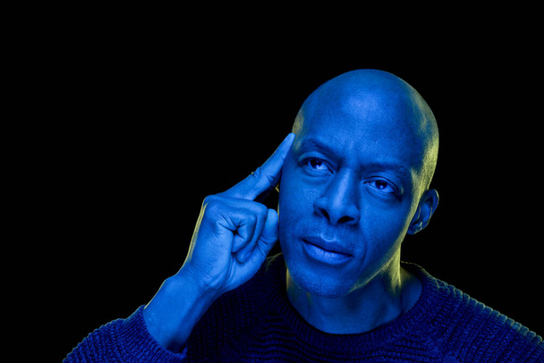 Φωτογραφία στούντιο με μπλε φως. Μαύρος άντρας με στοχαστική έκφραση, απομονωμένος σε μαύρο φόντο. Οριζόντια με copyspace - Φωτογραφία, εικόνα