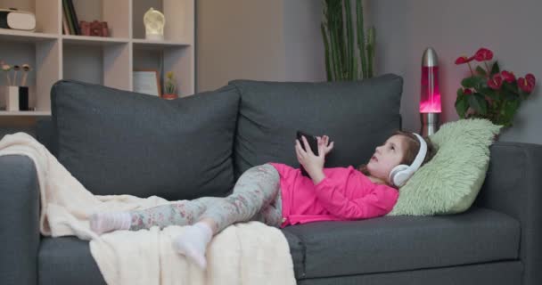 Happy Little girl in wireless headphones chilling laughing on sofa listening to favorite music holding phone using mobile online player app enjoy peaceful mood wearing earphones at home - Záběry, video