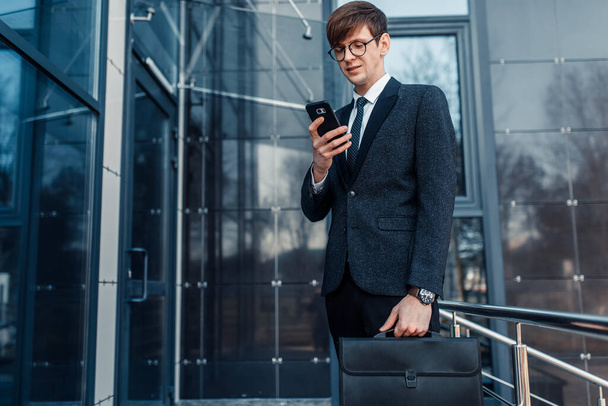 businessman in business suit holding his briefcase using his phone and standing on the street against the background of a glass business center - Zdjęcie, obraz