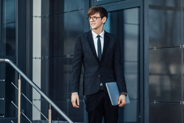 Attractive serious young businessman in a business suit, holding his laptop, outdoors on the background of a glass office - Фото, зображення