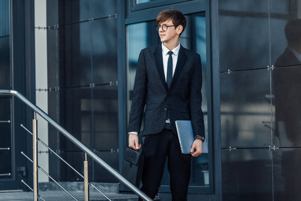 Attractive serious young businessman in a business suit, holding his laptop, outdoors on the background of a glass office - Zdjęcie, obraz