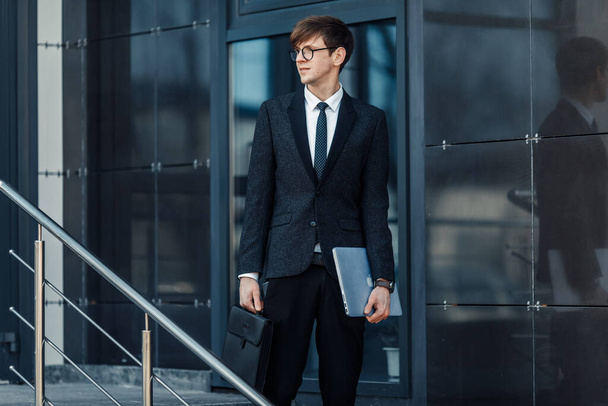 Atractivo joven empresario serio en un traje de negocios, sosteniendo su computadora portátil, al aire libre en el fondo de una oficina de vidrio
 - Foto, Imagen