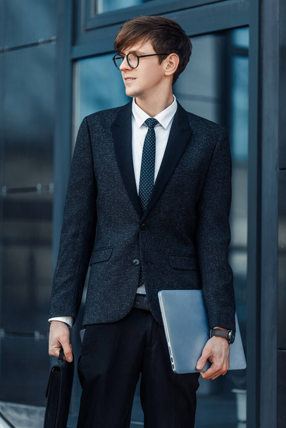 Attractive serious young businessman in a business suit, holding his laptop, outdoors on the background of a glass office - Foto, imagen