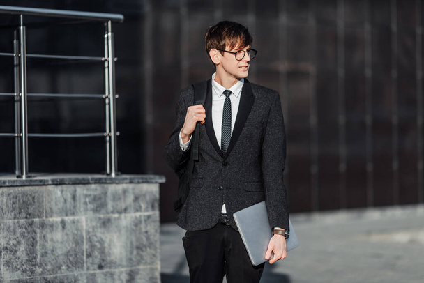 Attractive serious young businessman in a business suit, holding his laptop, outdoors on the background of a glass office - Фото, изображение