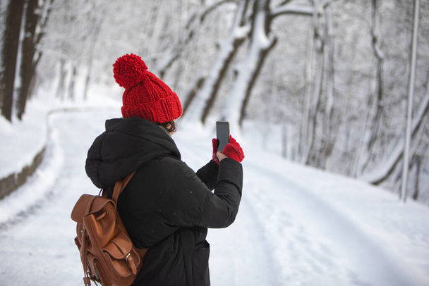 vrouw in de winter outfit wandelen door besneeuwde park weg het nemen van foto op telefoon - Foto, afbeelding