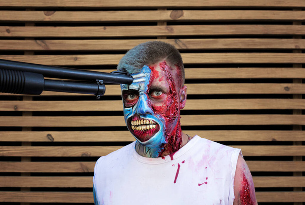 Jongeman in de make-up. Een zombie. Verscheurde wonden. op houten ondergrond. Halloween concept. gevangen zombies dreigen met een pistool tegen zijn hoofd - Foto, afbeelding