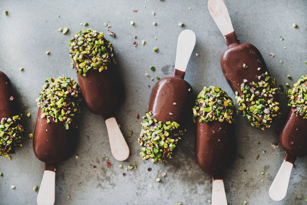Ice cream popsicle pattern. Flat-lay of chocolate glazed ice cream pops with pistachio icing over grey concrete background, top view, close-up. Summer seasonal cold sweet healthy vegan dessert - Photo, Image