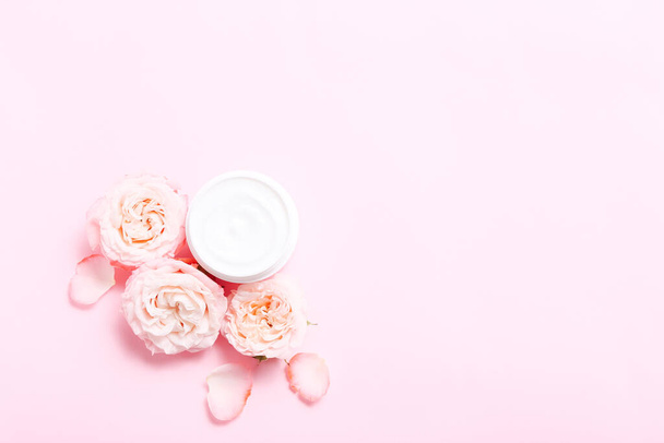 Open jar with cream for skin care and roses flowers around on pink background. - Photo, Image