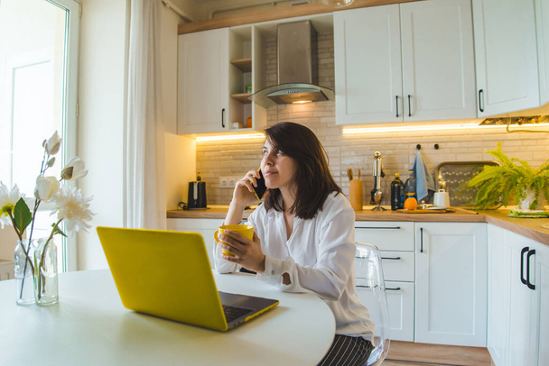 mujer sentada en la cocina trabajando en el portátil hablando por teléfono bebiendo té. estilo de vida doméstico
 - Foto, imagen