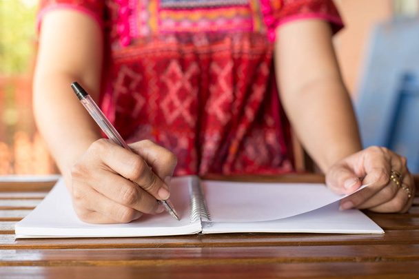 Kobieta trzymająca długopis pisze na notatniku i kubku na drewnianym stole z ciepłym światłem - Zdjęcie, obraz