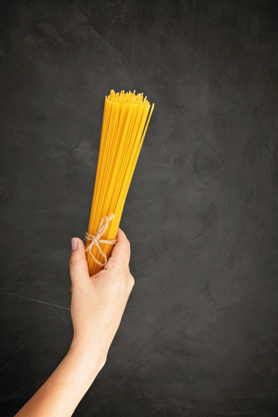 暗い背景にイタリアのパスタを調理するためのスパゲティを保持する女性の手のフラットレイアウト.伝統的なイタリア料理のコンセプトのトップビュー - 写真・画像