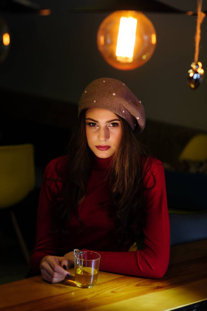 Retrato de una hermosa chica morena en un suéter rojo en la mesa con una taza de té en el crepúsculo
 - Foto, imagen