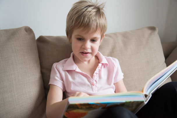 A child with short hair and blond hair, reading a large book. A schoolboy, a schoolgirl, snuggled up on the sofa. - Foto, afbeelding