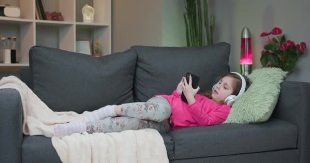 Little girl in wireless headphones chilling laughing on sofa listening to favorite music holding phone using mobile online player app. Enjoy peaceful mood wearing earphones at home - Filmati, video