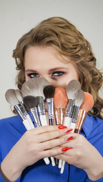 Young blonde girl hides her lips behind makeup brushes. The face is hidden behind curls of hair. Clearly expressed emotions on the womans face. - Fotoğraf, Görsel