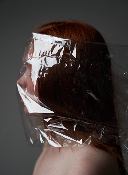Πορτρέτο μιας νεαρής κοκκινομάλλας σε γκρι φόντο με μια πλαστική σακούλα στο κεφάλι της. Η έννοια της πλαστικής ρύπανσης της φύσης. Η περίσσεια πλαστικού στη ζωή ενός ατόμου                                            - Φωτογραφία, εικόνα