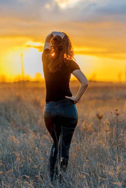 Mujer en pantalones y una camiseta posando sobre un fondo al atardecer, vista trasera. Belleza de la naturaleza
. - Foto, Imagen