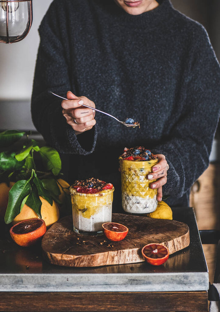 Young woman eating overnight oat bircher muesli with mango smoothie, buckwheat granola and fresh ripe berries from glass with spoon over grey concrete kitchen counter. Healthy, clean eating concept - Fotó, kép