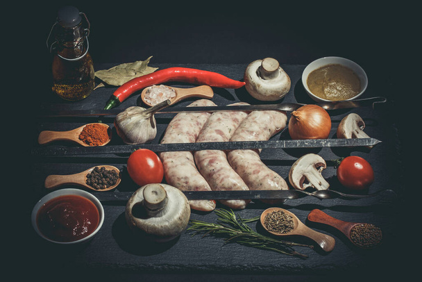 Raw sausages with skewers, different vegetables, spices and ingredients on a black background - Фото, изображение