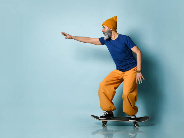 Ηλικιωμένος άντρας με μπλουζάκι, πορτοκαλί παντελόνι, καπέλο, παπούτσια. Ιππασία μαύρο skateboard ποζάρουν πλάγια σε μπλε φόντο. Πλήρες μήκος - Φωτογραφία, εικόνα