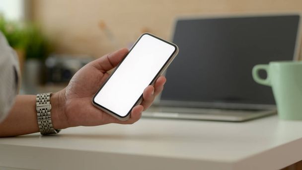 Cropped shot of male freelancer holding blank screen smartphone while sitting in simple working space - Photo, Image