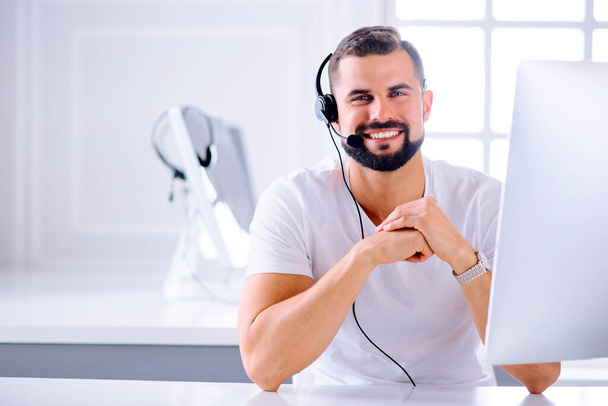 Młody uśmiechnięty operator obsługi klienta z słuchawkami głośnomówiącymi pracującymi w biurze. Pojęcie - Zdjęcie, obraz