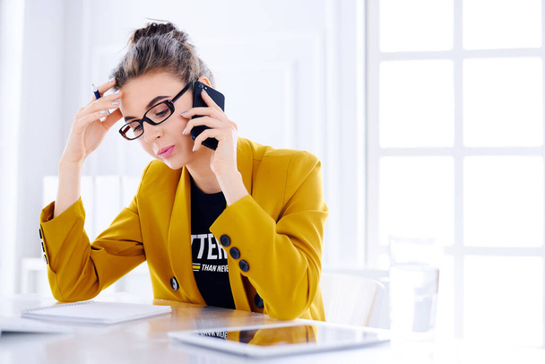 Νεαρή επιχειρηματίας μιλάει στο τηλέφωνο στο γραφείο της και κρατάει το κεφάλι της - Φωτογραφία, εικόνα