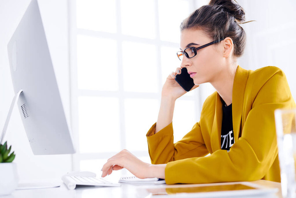 Молодая деловая женщина разговаривает по телефону во время использования рабочего стола
 - Фото, изображение