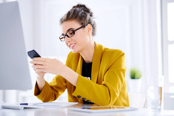 İş kadını, bilgisayarın önünde masada otururken telefonuyla bir mesaj yazar. - Fotoğraf, Görsel