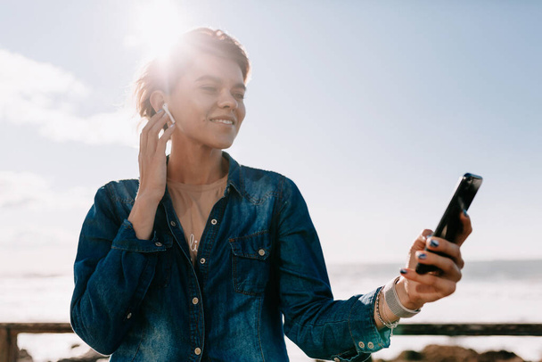 Draußen Porträt einer glücklich lächelnden Frau im Jeanshemd mit Smartphone, die im Sonnenschein am Meer Musik hört. - Foto, Bild