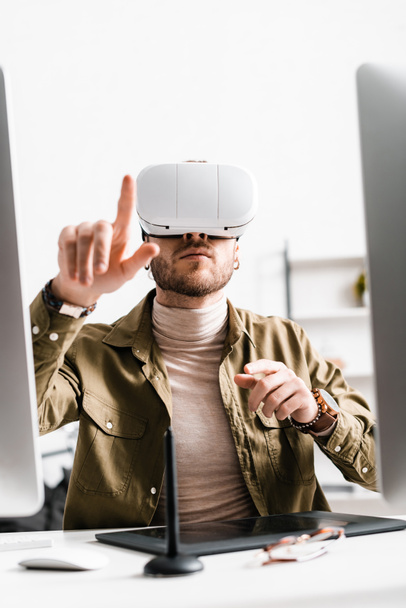 Selectieve focus van digitale ontwerper in virtual reality headset wijzend met vinger in de buurt van grafische tablet en computers op tafel - Foto, afbeelding