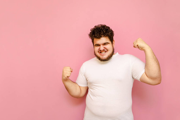 Портрет радісного чоловіка в білій футболці з надмірною вагою радіє перемозі на рожевому тлі, заглядає в камеру з щасливим обличчям. Веселий радісний товстун підняв руки вгору, ізольований
. - Фото, зображення