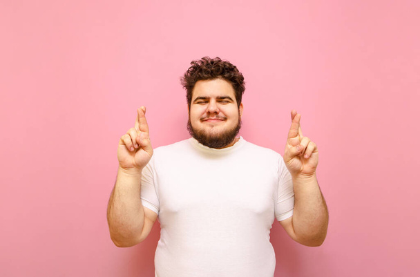 Happy σγουρά λίπος άνθρωπος στέκεται σε ένα ροζ φόντο και κάνει μια ευχή με σταυρωμένα δάχτυλα σε ένα ροζ φόντο. Χαρούμενο υπέρβαρο αγόρι σταυρώνει τα δάχτυλα με κλειστά μάτια, φορώντας λευκό μπλουζάκι, απομονωμένο. - Φωτογραφία, εικόνα