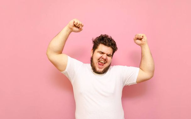 Happy fat young man shouting with joy with his hands raised on a pink background. Joyful overweight guy and white T-shirt, emotionally happy to win. Winning concept. - Photo, image
