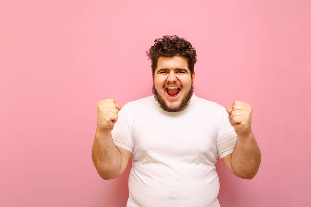 Retrato de un chico alegre con una camiseta blanca y sobrepeso regocija la victoria de su amado equipo sobre un fondo rosa. Feliz hombre alegre y gordo con barba levantó las manos de alegría. Copiar espacio
 - Foto, imagen