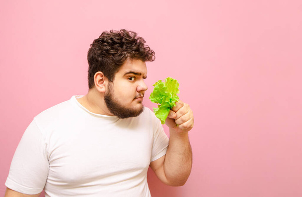 Смешной толстяк в белой футболке стоит на розовом фоне с салатом в руках, нюхает свежую зелень и подозрительно смотрит в камеру одним глазом. Изолирована. Толстый смешной человек на диете
. - Фото, изображение