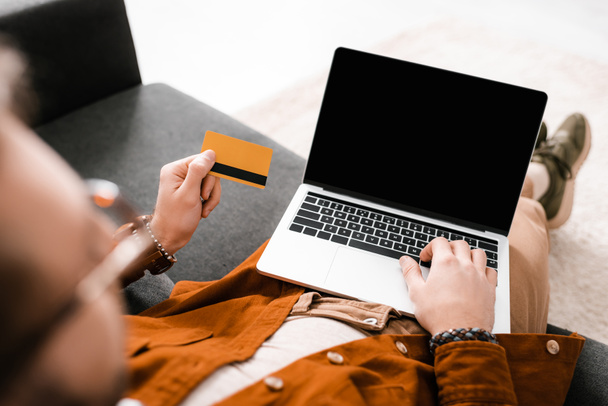 Selective focus of digital designer using laptop with blank screen and holding credit card on couch - Fotoğraf, Görsel