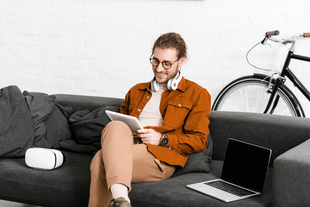 Smiling digital artist using tablet near vr headset and laptop on couch  - Φωτογραφία, εικόνα