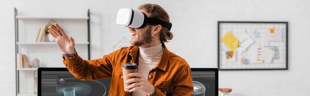 Panoramic shot of cheerful 3d artist in vr headset holding coffee to go near project of 3d design on computer monitors in office - Photo, Image