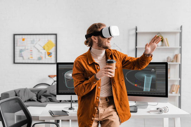 Cheerful digital designer in virtual reality headset holding coffee to go near 3d design project on computer monitors on table - Φωτογραφία, εικόνα
