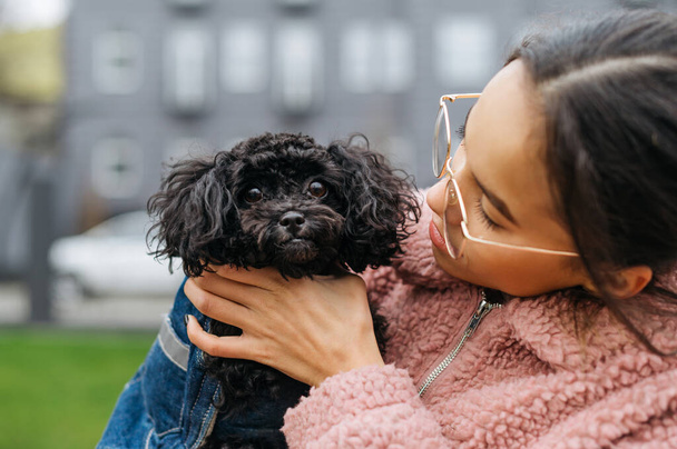 Closeup photo of a cute dog breed toy poodle in the hands of an attractive girl. A woman in glasses and a pink coat holds a funny puppy in her hand. Owner walks with her pet on the streets. - Photo, Image