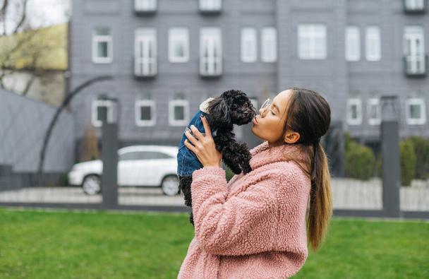 Beautiful girl owner in pink coat holds black curly puppy in her hands and kisses, doggy looks into the camera. Walk with your pet on a cool day. Lady with puppy in her arms walking down the street. - Photo, Image