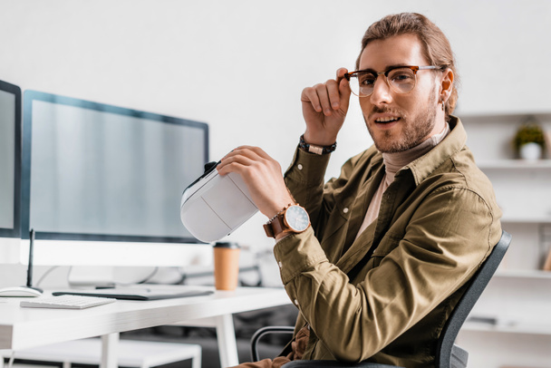 Hezký 3D umělec drží vr sluchátka a dívá se na fotoaparát u stolu v kanceláři  - Fotografie, Obrázek