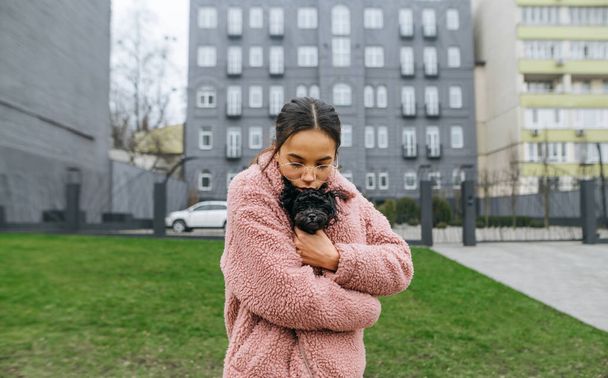 Nette Frau mit Brille und rosa Mantel steht auf der Straße, einen Welpen im Arm, umarmt und küsst ein Haustier. Attraktive Dame küsst Hundepudel auf einem Spaziergang. Liebe und Haustiere - Foto, Bild