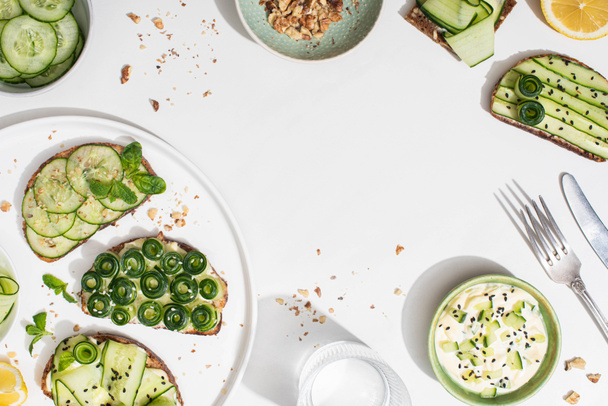 top view of fresh cucumber toasts on plate near lemon, water, cutlery and yogurt on white background - Foto, Bild