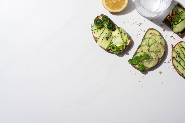 top view of fresh cucumber toasts with seeds, mint and basil leaves, lemon near water on white background - Foto, Bild