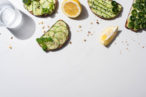 top view of fresh cucumber toasts with seeds, mint and basil leaves, lemon near water on white background - Foto, imagen
